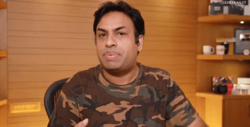 A Man Wearing A Camouflage Shirt Is Sitting In Front Of A Shelf With Geekkiranjit Written On It GIF