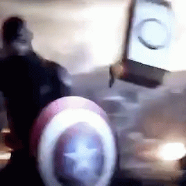 A Man Wearing A Captain America Shield Is Standing In A Dark Room . GIF