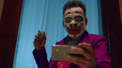 A Man Wearing A Clown Mask Is Holding A Rock And Looking At A Cell Phone GIF