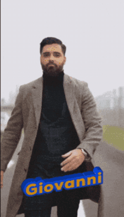 A Man Wearing A Coat And A Black Turtleneck Has The Name Giovanni Written On A Blue Sign GIF