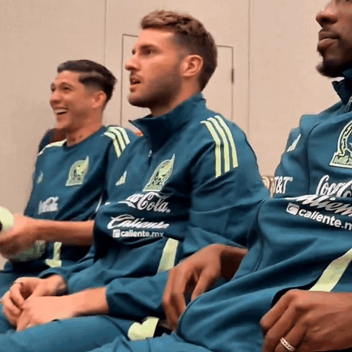 A Man Wearing A Coca Cola Shirt Sits Next To Another Man Wearing A Caliente Shirt GIF