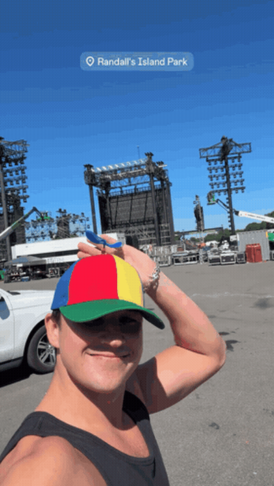 A Man Wearing A Colorful Hat Is Standing In Front Of A Large Stage At Randall 'S Island Park GIF