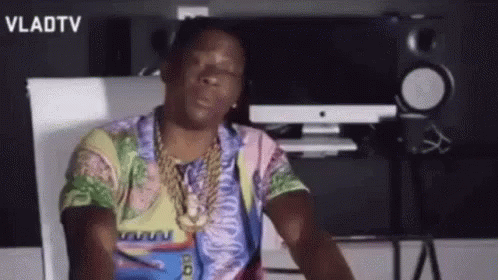 A Man Wearing A Colorful Shirt And A Gold Chain Is Sitting In Front Of A Computer . GIF