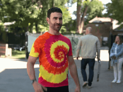 A Man Wearing A Colorful Tie Dye Shirt Is Walking Down A Sidewalk GIF