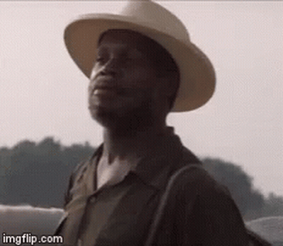 A Man Wearing A Cowboy Hat And A Backpack Is Standing In Front Of A Body Of Water . GIF