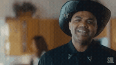 A Man Wearing A Cowboy Hat And A Black Shirt Is Smiling And Looking At The Camera . GIF