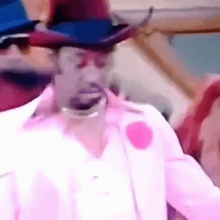 A Man Wearing A Cowboy Hat And A Pink Shirt Is Looking At The Camera . GIF