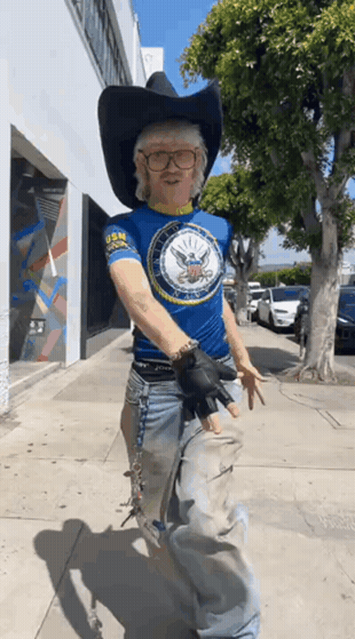 A Man Wearing A Cowboy Hat And Glasses Is Walking Down The Street . GIF