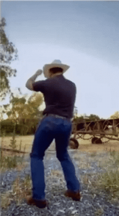 A Man Wearing A Cowboy Hat And Jeans Is Standing In A Field . GIF