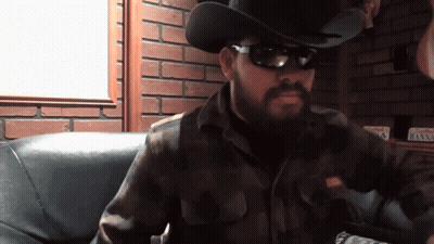 A Man Wearing A Cowboy Hat And Sunglasses Is Sitting In A Chair GIF