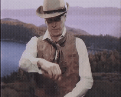 A Man Wearing A Cowboy Hat And Vest Looks At His Watch GIF