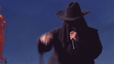 A Man Wearing A Cowboy Hat Covering His Face Holds A Microphone GIF
