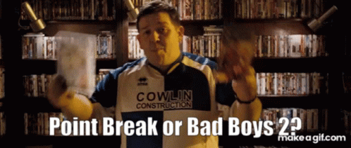 A Man Wearing A Cowlin Construction Shirt Is Standing In Front Of A Bookshelf GIF