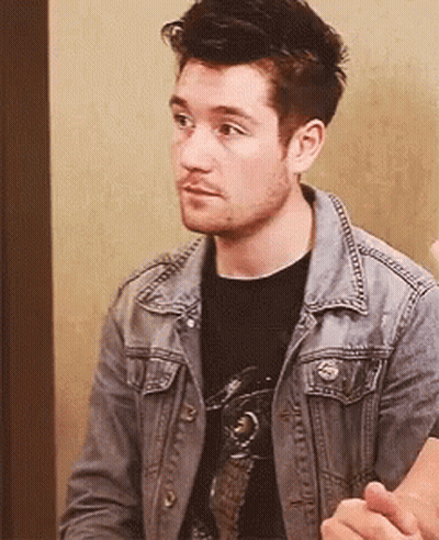 A Man Wearing A Denim Jacket And A Black Shirt Is Sitting In A Chair . GIF