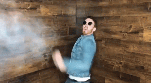 A Man Wearing A Denim Jacket And Sunglasses Is Dancing In Front Of A Wooden Wall GIF