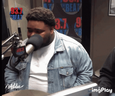 A Man Wearing A Denim Jacket Is Speaking Into A Microphone In Front Of A Sign That Says 97.7 The Beat GIF