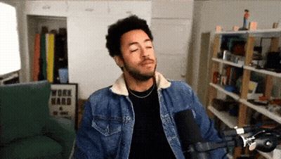 A Man Wearing A Denim Jacket Stands In Front Of A Microphone With A Work Hard Poster On The Wall Behind Him GIF