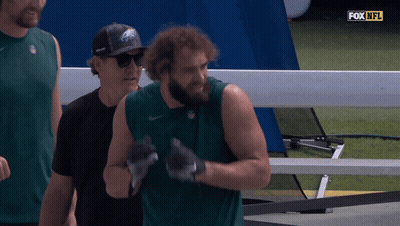 A Man Wearing A Eagles Hat Stands Next To A Man Wearing A Black Shirt GIF