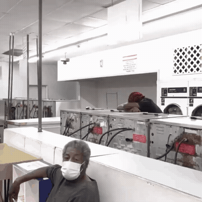 A Man Wearing A Face Mask Sits In A Laundromat Surrounded By Washers And Dryers GIF