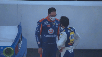 A Man Wearing A Face Mask Talks To Another Man Wearing A Nascar Uniform GIF