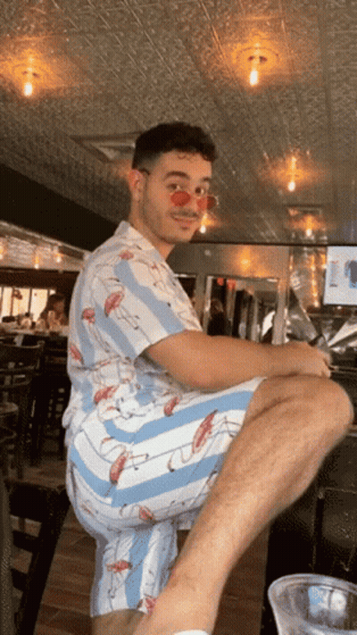A Man Wearing A Flamingo Shirt And Shorts Sits On A Bar Stool GIF