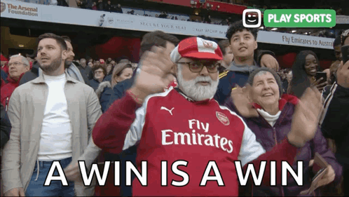 A Man Wearing A Fly Emirates Jersey Stands In A Crowd Of People GIF