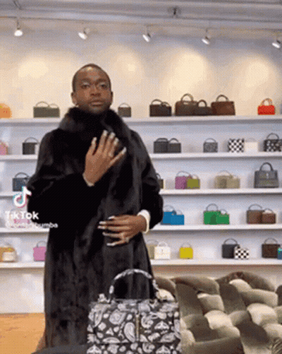 A Man Wearing A Fur Coat Is Standing In Front Of A Wall Of Bags . GIF