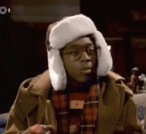 A Man Wearing A Fur Hat And Glasses Is Sitting Down . GIF