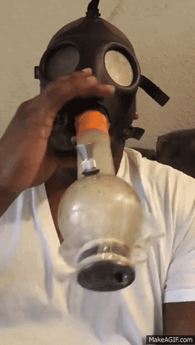 A Man Wearing A Gas Mask Is Blowing Smoke Out Of A Bong GIF