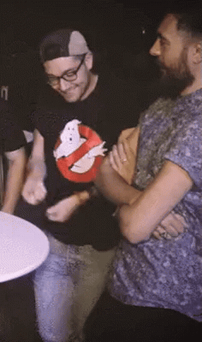 A Man Wearing A Ghostbusters Shirt Is Standing Next To Another Man With His Arms Crossed GIF