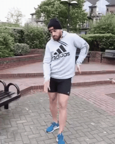 A Man Wearing A Gray Adidas Hoodie And Black Shorts Is Standing On A Brick Sidewalk . GIF