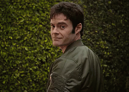 A Man Wearing A Green Bomber Jacket Is Standing In Front Of A Bush . GIF