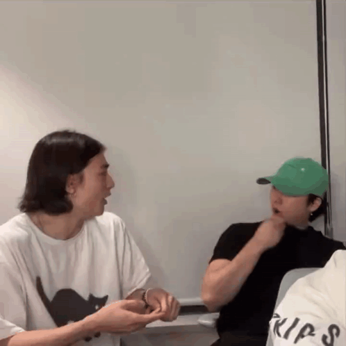 A Man Wearing A Green Hat Is Sitting Next To Another Man Wearing A White Shirt With A Black Cat On It . GIF