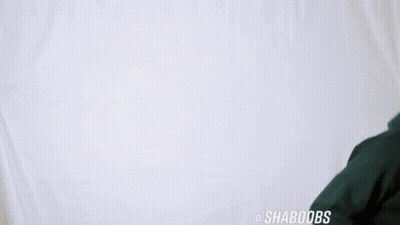 A Man Wearing A Green Hoodie Is Dancing In Front Of A White Wall . GIF
