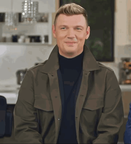 A Man Wearing A Green Jacket And A Black Turtleneck Smiles For The Camera GIF