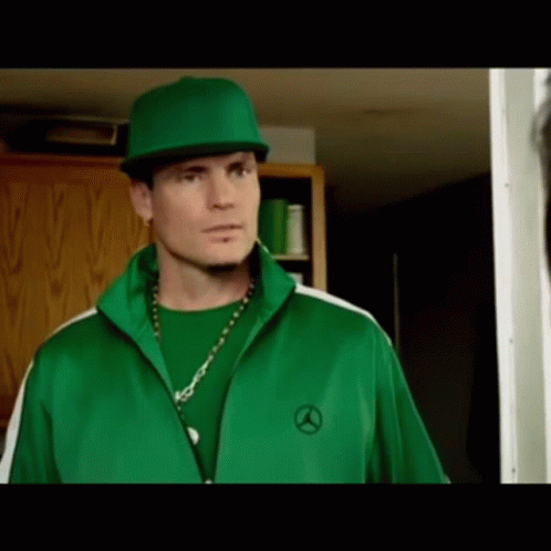 A Man Wearing A Green Jacket And A Green Hat Is Standing In A Doorway . GIF