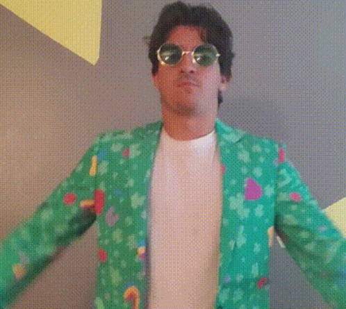 A Man Wearing A Green Jacket And Sunglasses Stands In Front Of A Wall GIF