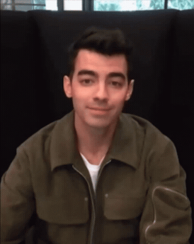 A Man Wearing A Green Jacket Is Sitting In A Chair And Smiling GIF