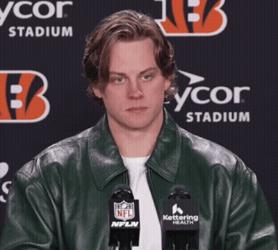 A Man Wearing A Green Leather Jacket Stands In Front Of Two Microphones With A Nfl Logo On Them GIF