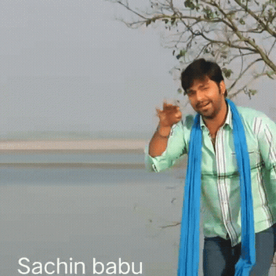 A Man Wearing A Green Shirt And A Blue Scarf Stands In Front Of A Body Of Water GIF