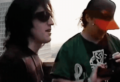 A Man Wearing A Green Shirt With The Letter L On It Stands Next To Another Man Wearing An Orange Hat GIF