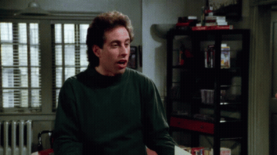 A Man Wearing A Green Sweater Is Sitting In Front Of A Shelf With A Box That Says Dominoes GIF