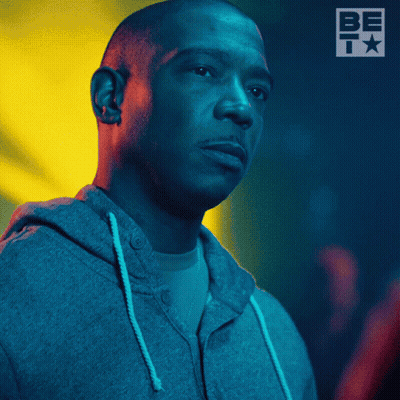 A Man Wearing A Grey Hoodie Stands In Front Of A Sign That Says Be T GIF