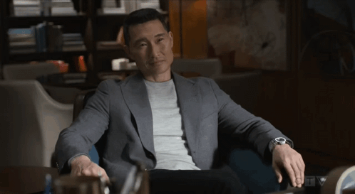 A Man Wearing A Grey Jacket And A Watch Sits In A Dark Room GIF