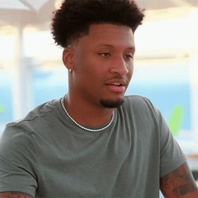A Man Wearing A Grey Shirt And A Pearl Necklace Looks At The Camera GIF