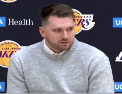 A Man Wearing A Grey Sweater Stands In Front Of A Los Angeles Lakers Logo GIF