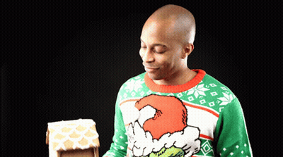 A Man Wearing A Grinch Sweater Looks At A Gingerbread House GIF