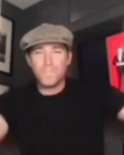 A Man Wearing A Hat And A Black Shirt Is Standing In A Room . GIF