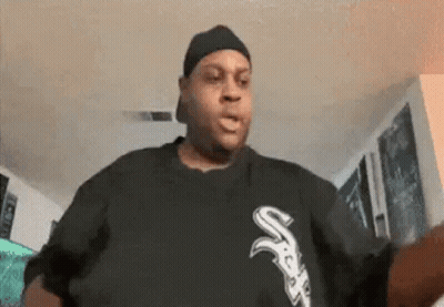 a man wearing a hat and a black shirt with the letter s on it is standing in a living room .