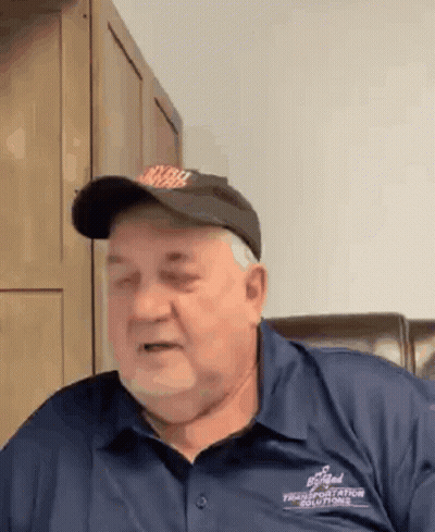 A Man Wearing A Hat And A Blue Shirt Is Sitting In A Chair And Making A Funny Face . GIF